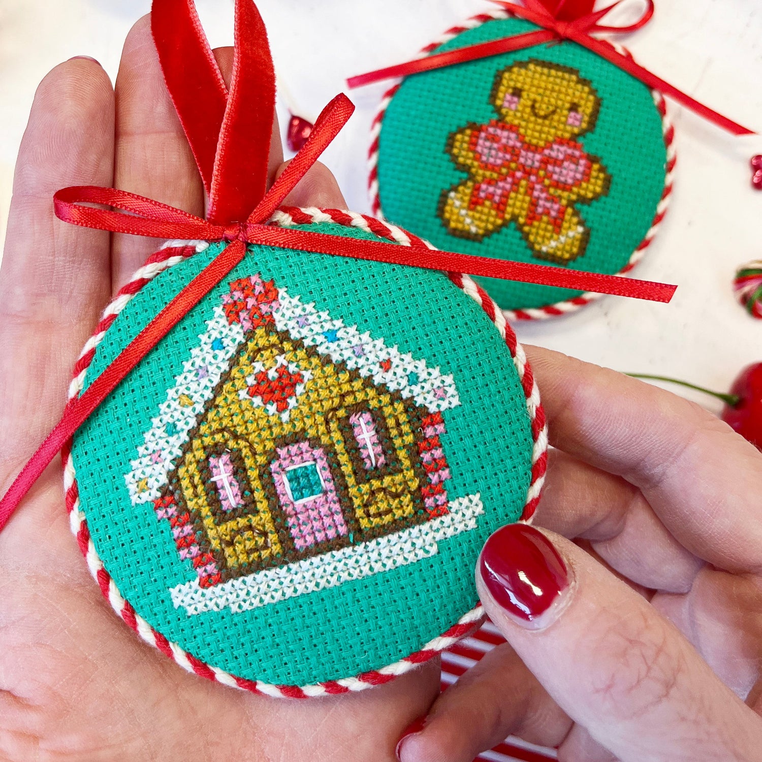 Hand holding a cross-stitched Christmas ornament with a gingerbread design, surrounded by festive decorations.