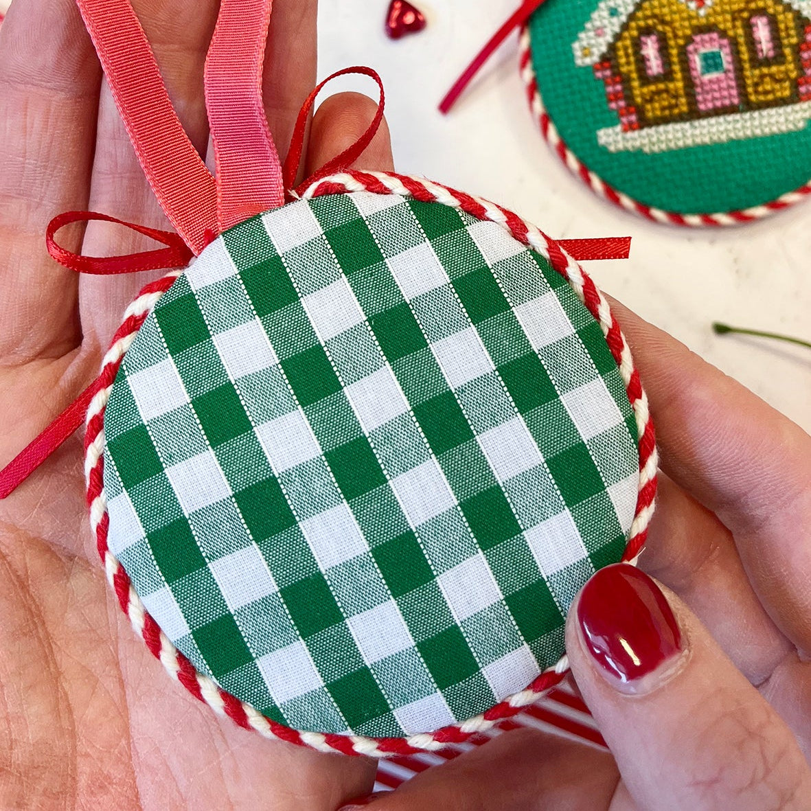 Green and white gingham ornament with red ribbon held by a hand, with other ornaments in the background.