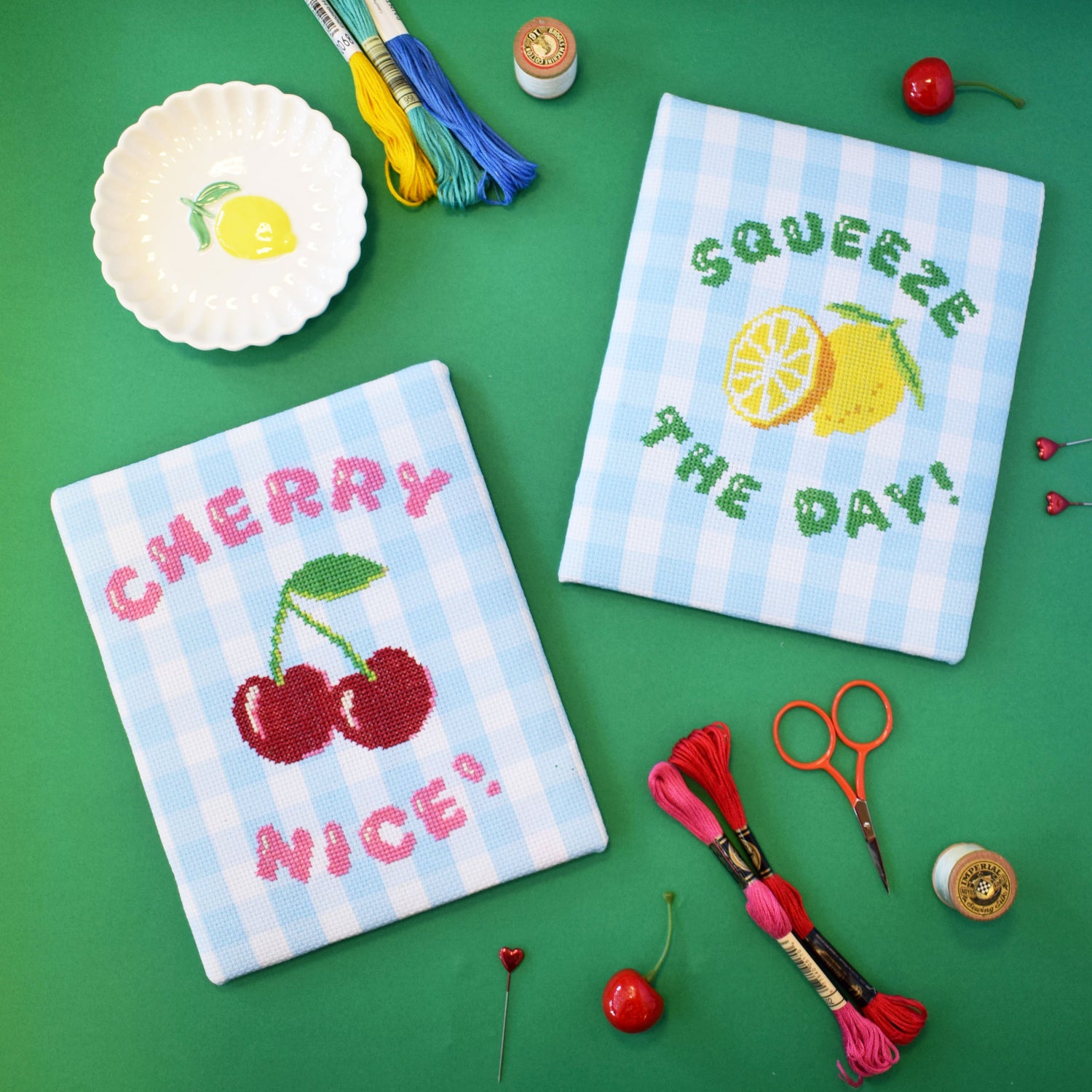 set of cross stitch designs with a 'squeeze the day' slogan and lemons motif and a 'cherry nice' design. The designs are stitched onto blue gingham and are on a green background.