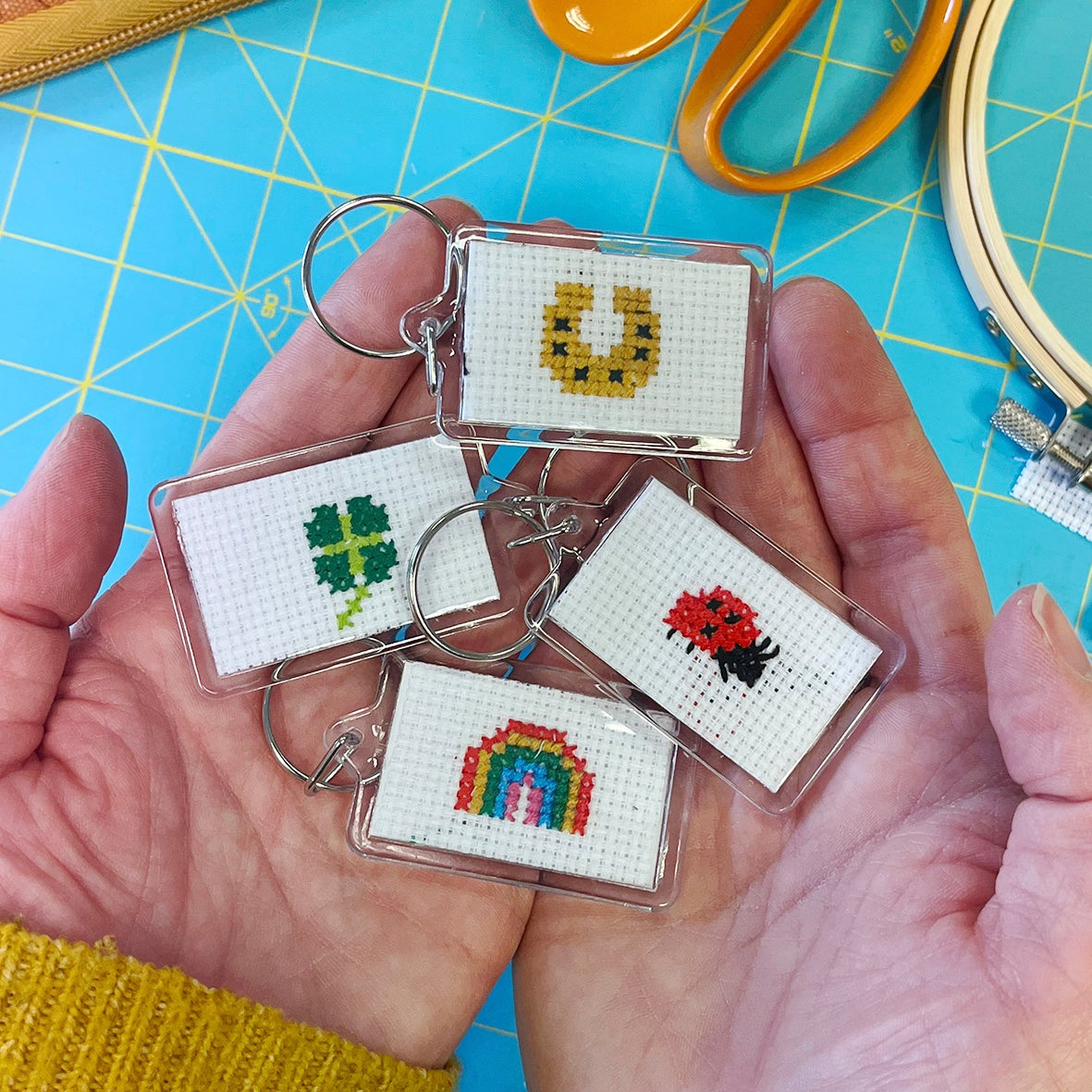 Handmade keychains with cross-stitch designs held by a person on a crafting table.