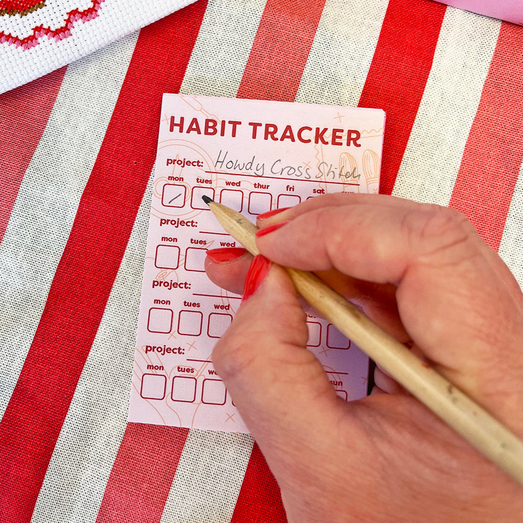 Habit tracker card on a red and white striped fabric with a hand holding a pen.