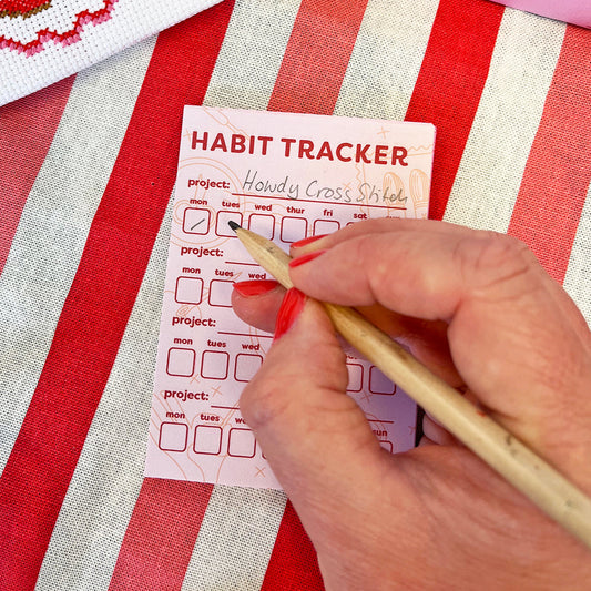 Habit tracker card on a red and white striped fabric with a hand holding a pen.