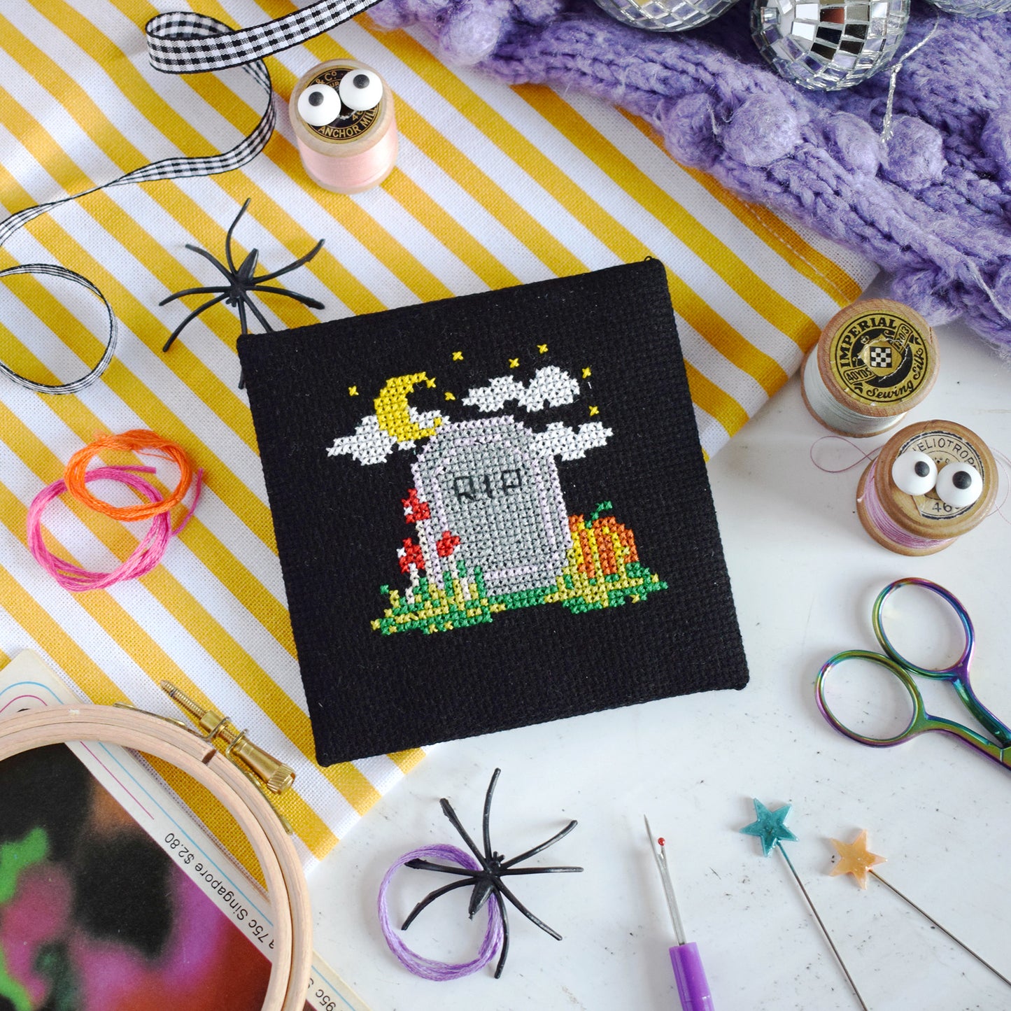 A finished black cross-stitch canvas featuring a Halloween tombstone design with a yellow crescent moon, white clouds, red flowers, and a pumpkin, styled in a flat-lay with craft supplies on a yellow-and-white striped fabric background.