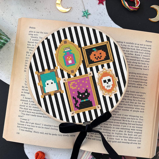 Decorative Halloween-themed cross stitch kit on black and white striped aida on an open book.
