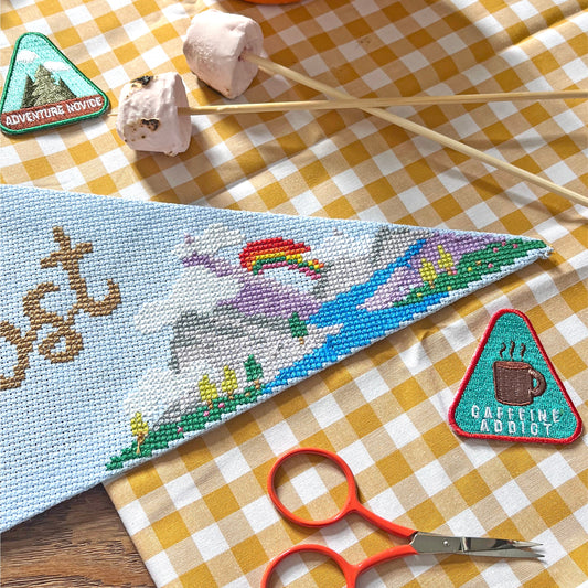 close up of a mountain themed design Cross-stitched onto a pennant with nature design, orange mug, scissors, and decorative elements on a checkered cloth