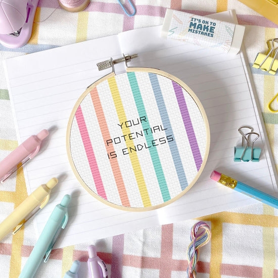 cross stitch hoop with pastel stripe rainbow pattern and text 'Your potential is endless' on a colourful fabric background.