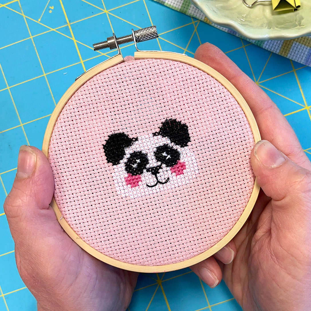 Cross-stitched panda design held in a wooden hoop on a blue cutting mat.