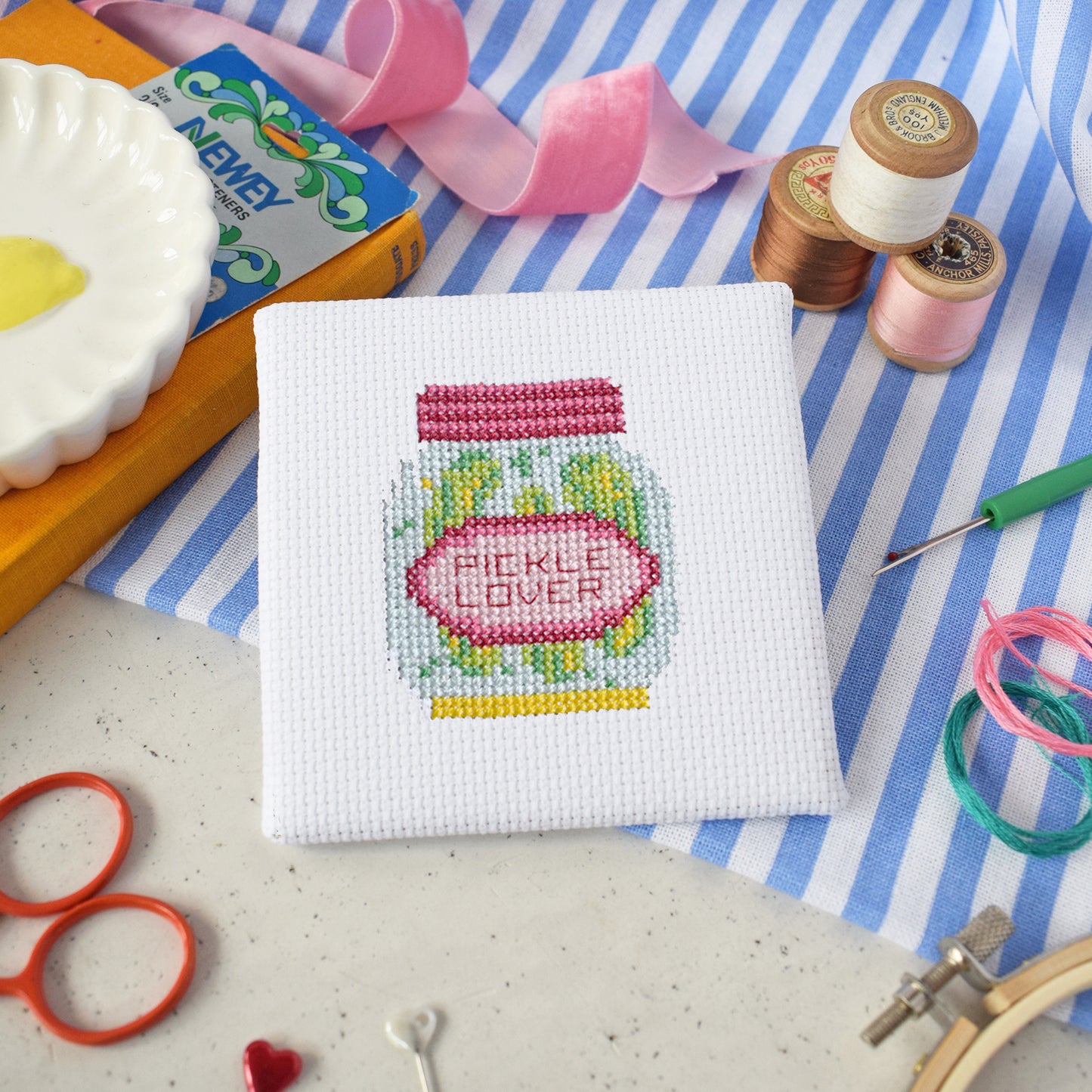 Cross-stitched pickle lover design on fabric with sewing supplies on a striped background