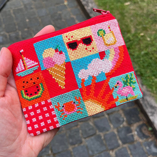 Hand holding a cross stitched red coin purse with colourful animal designs on a blurred outdoor background