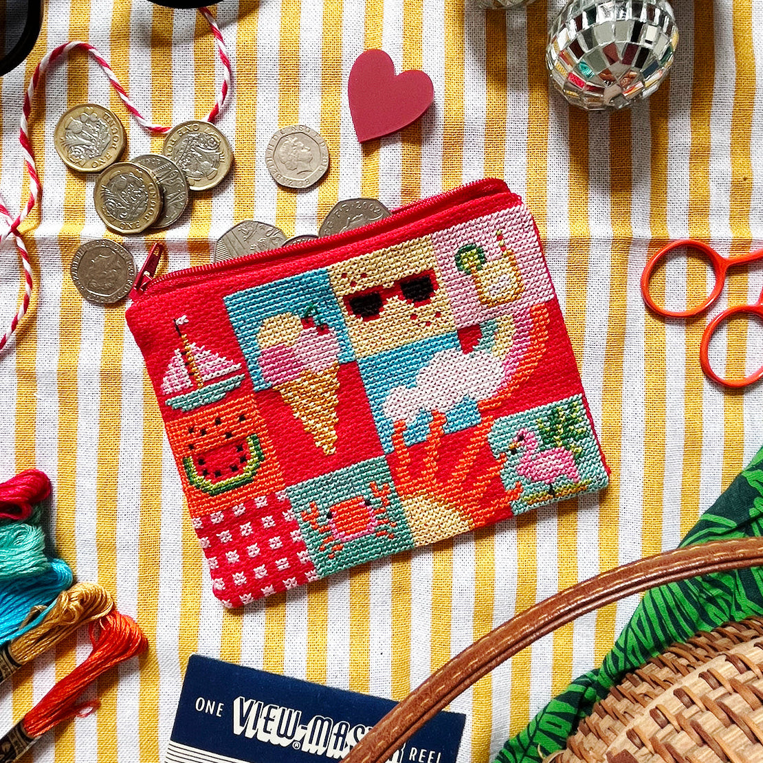 Colourful cross stitch purse with a beach theme and sewing supplies on a striped fabric background