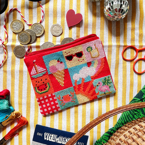 Colourful cross stitch purse with a beach theme and sewing supplies on a striped fabric background