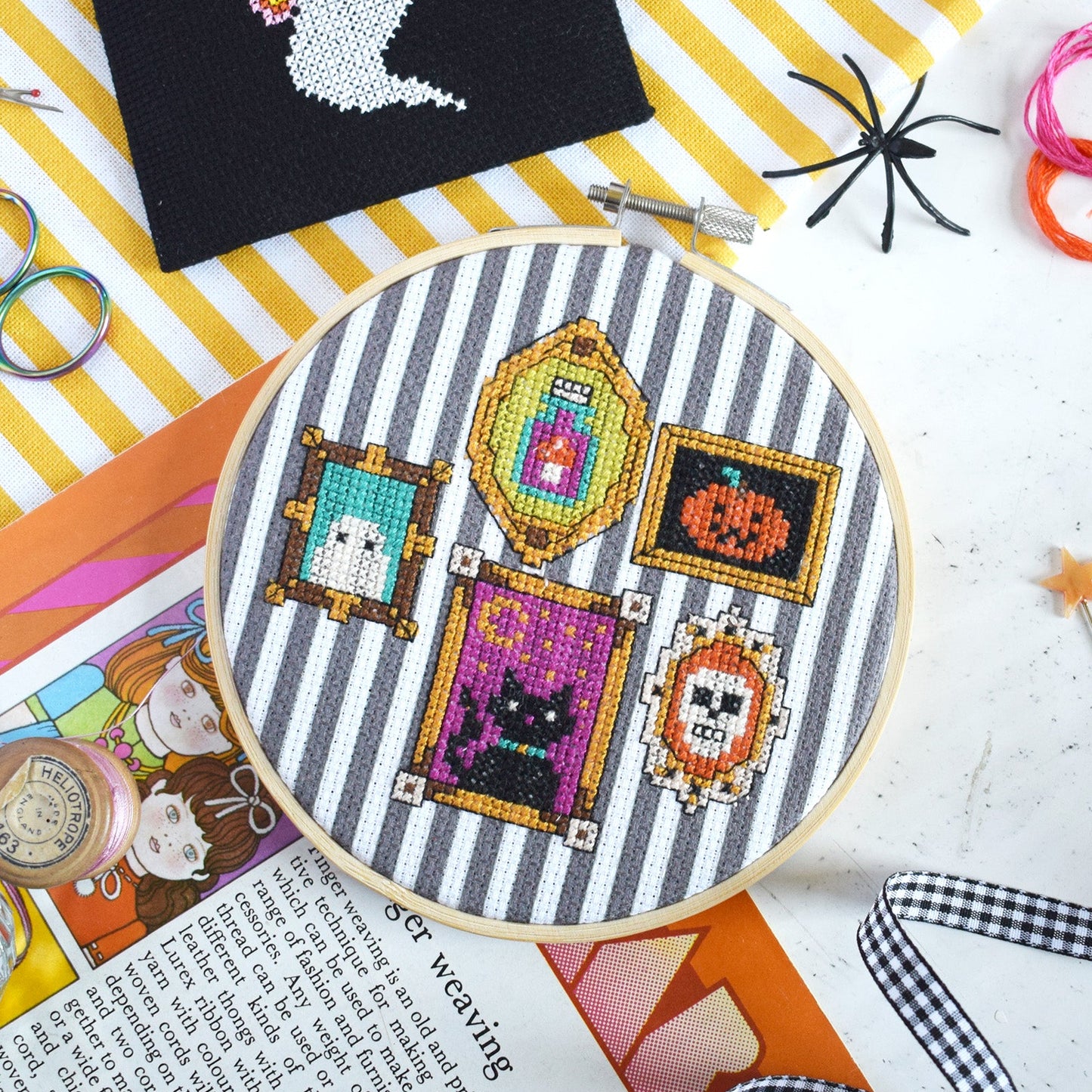 A wooden hoop with several colorful stitched frames including a cat, pumpkin, and moon on a black-and-white striped fabric. The scene is decorated with scissors, spider cutouts, a purple knit item, and other Halloween-themed craft props on a yellow-and-white striped surface.