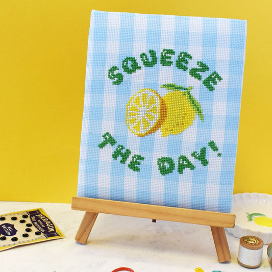 cross stitch kit with a 'squeeze the day' slogan and lemons motif. The design is stitched onto blue gingham and project is on a small easel against a bright yellow background
