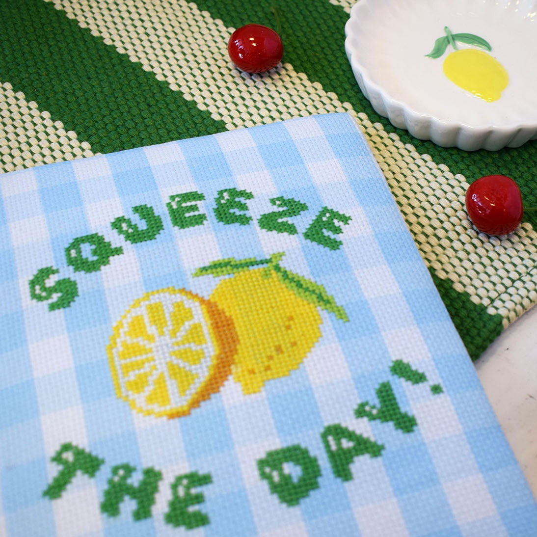 cross stitch kit with a 'squeeze the day' slogan and lemons motif. The design is stitched onto blue gingham and the design is displayed on a green and white background with a cute lemon trinket dish and cherries.