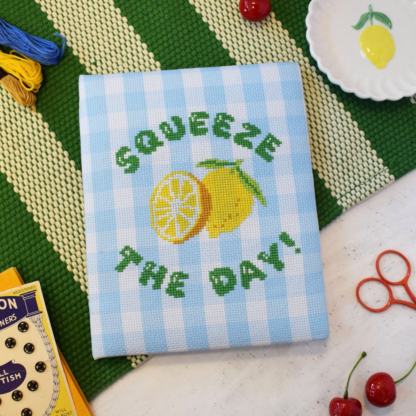cross stitch kit with a 'squeeze the day' slogan and lemons motif. The design is stitched onto blue gingham and the design is displayed on a green and white background with a cute lemon trinket dish.