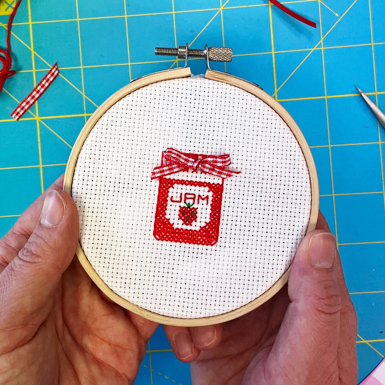 Cross-stitched design of a jar with 'Jam' text in a hoop on a blue cutting mat.