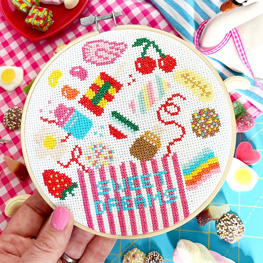 A hand holds a wooden embroidery hoop containing a colorful cross-stitch design of sweets and the words 'SWEET DREAMS' on white Aida fabric, surrounded by candy props on a blue cutting mat.